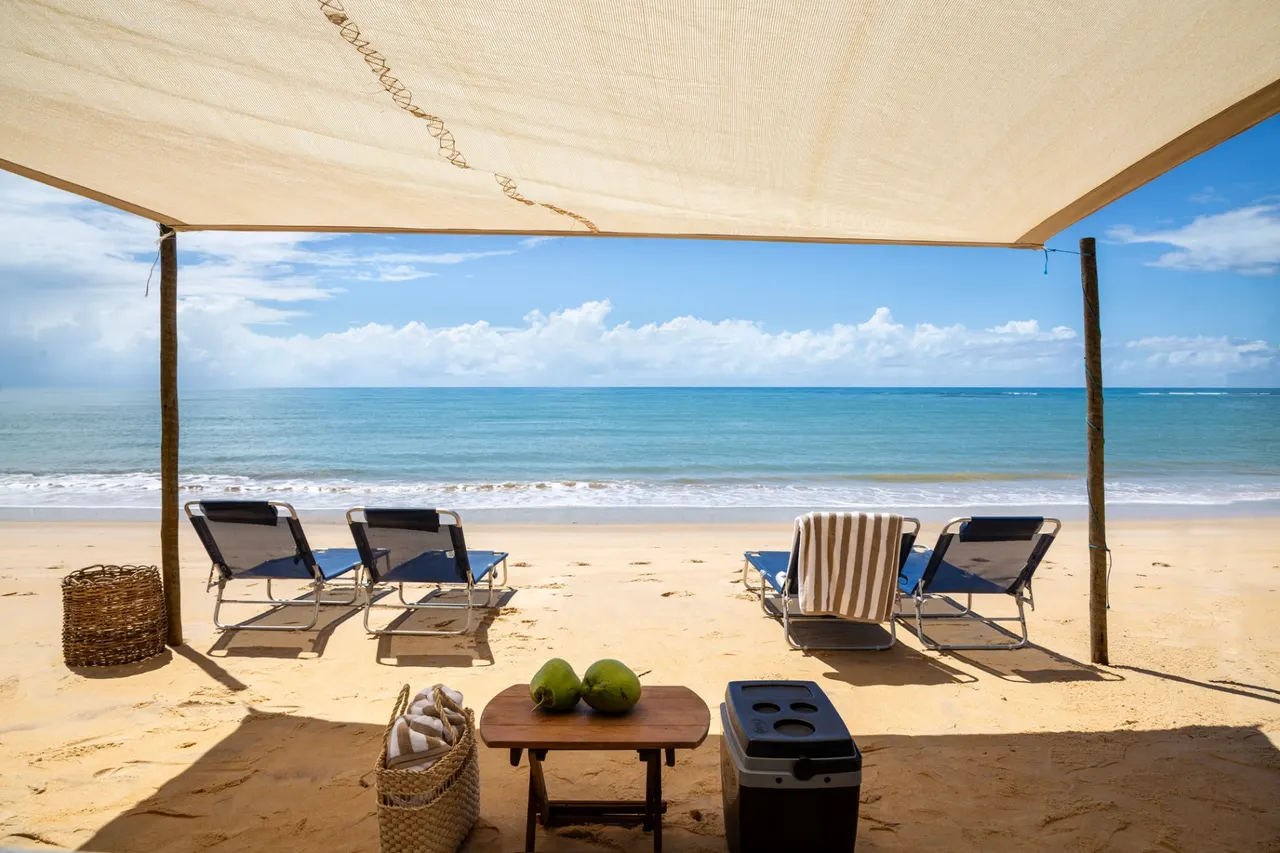 Tenda de praia com espreguiçadeiras, cadeiras e cooler diante do mar azul-turquesa em destino premium do litoral brasileiro
