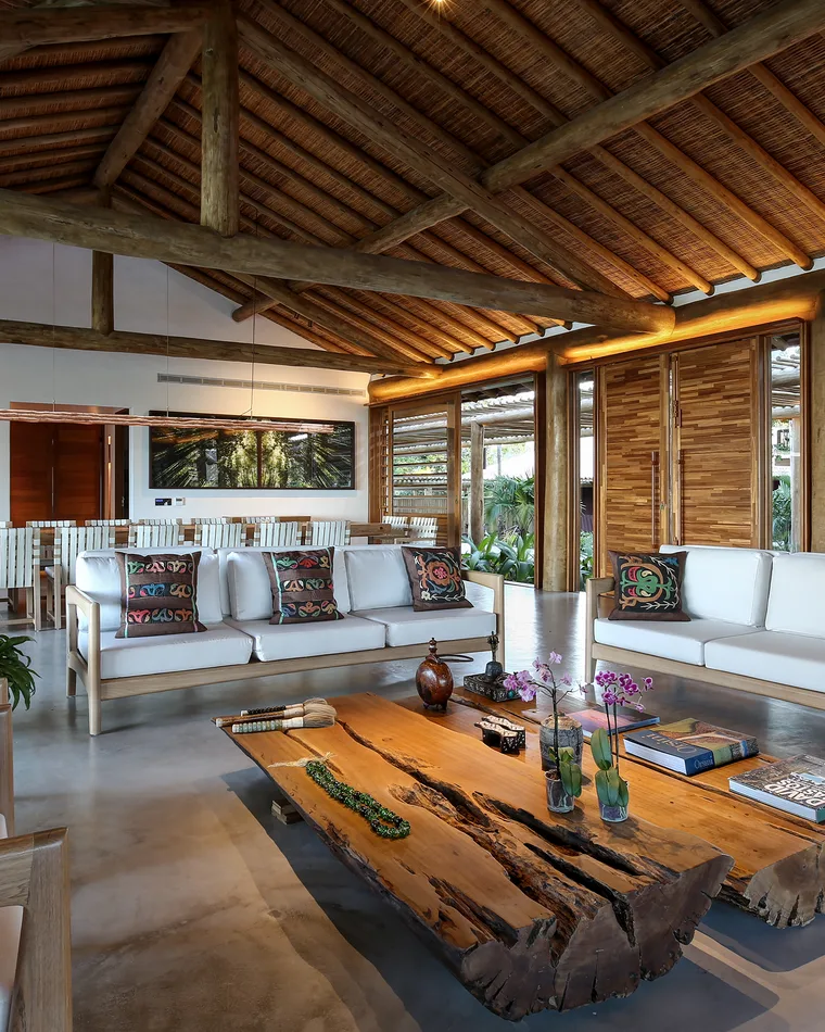 Sala de estar ampla de casa de alto padrão em Trancoso, com teto de palha em estrutura de madeira, sofás claros e mesa de centro rústica de madeira maciça
